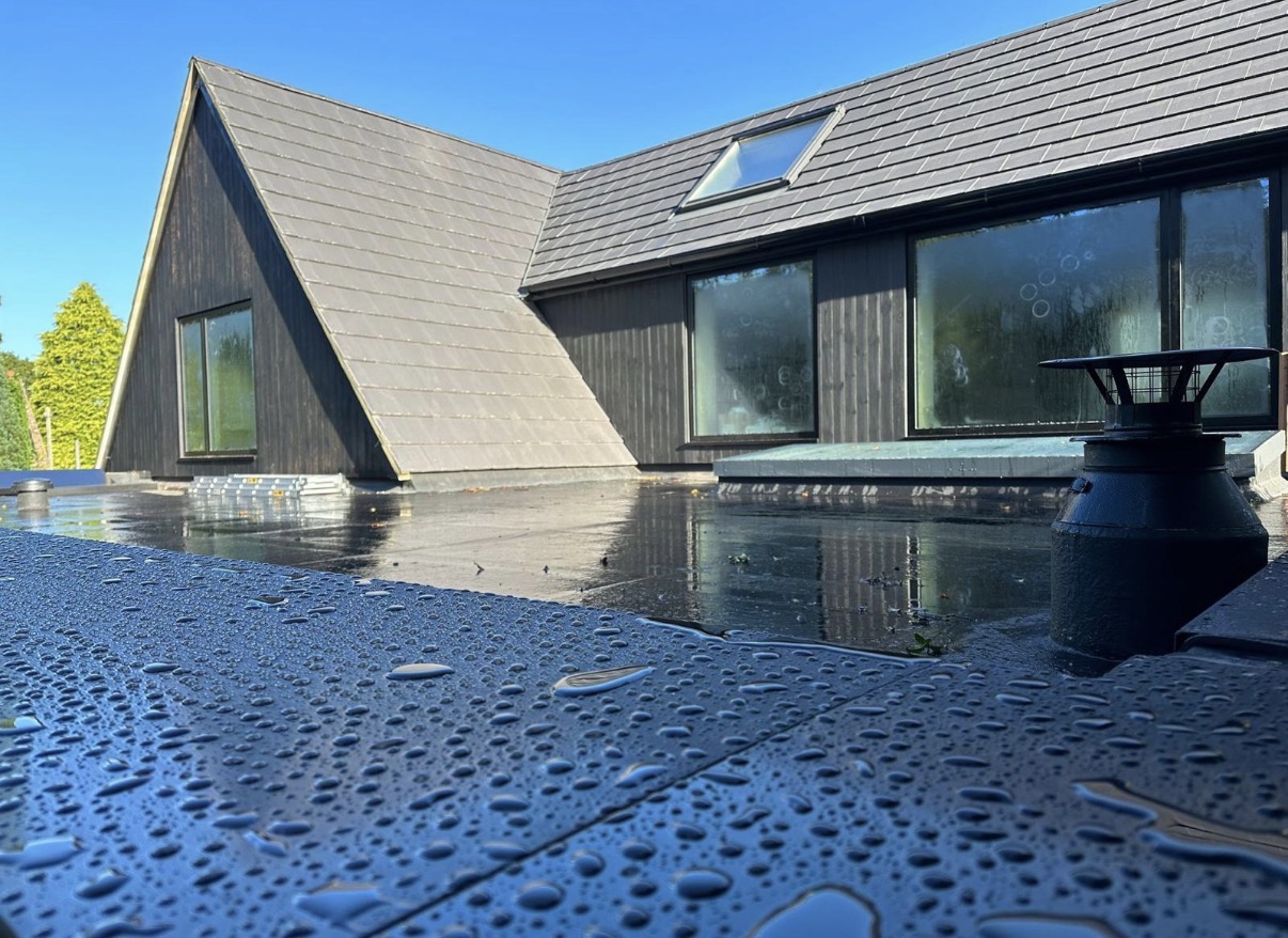Wet dark flat-roof surface with a vent, adjoining pitched roof and roof window